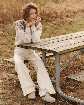 A woman sits at a wooden picnic table outside, wearing a fair isle sweater, cream-colored jeans, and clogs. She rests her chin on her hands.