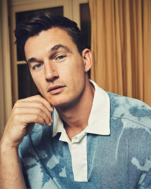 Portrait of an adult male with dark hair and blue eyes, resting his chin on his hand, wearing a blue acid-wash polo shirt indoors.
