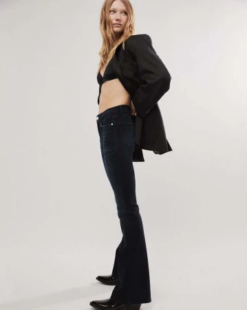 A young woman in a black bra, flare jeans, blazer, and pointed boots poses in a studio against a plain white backdrop.