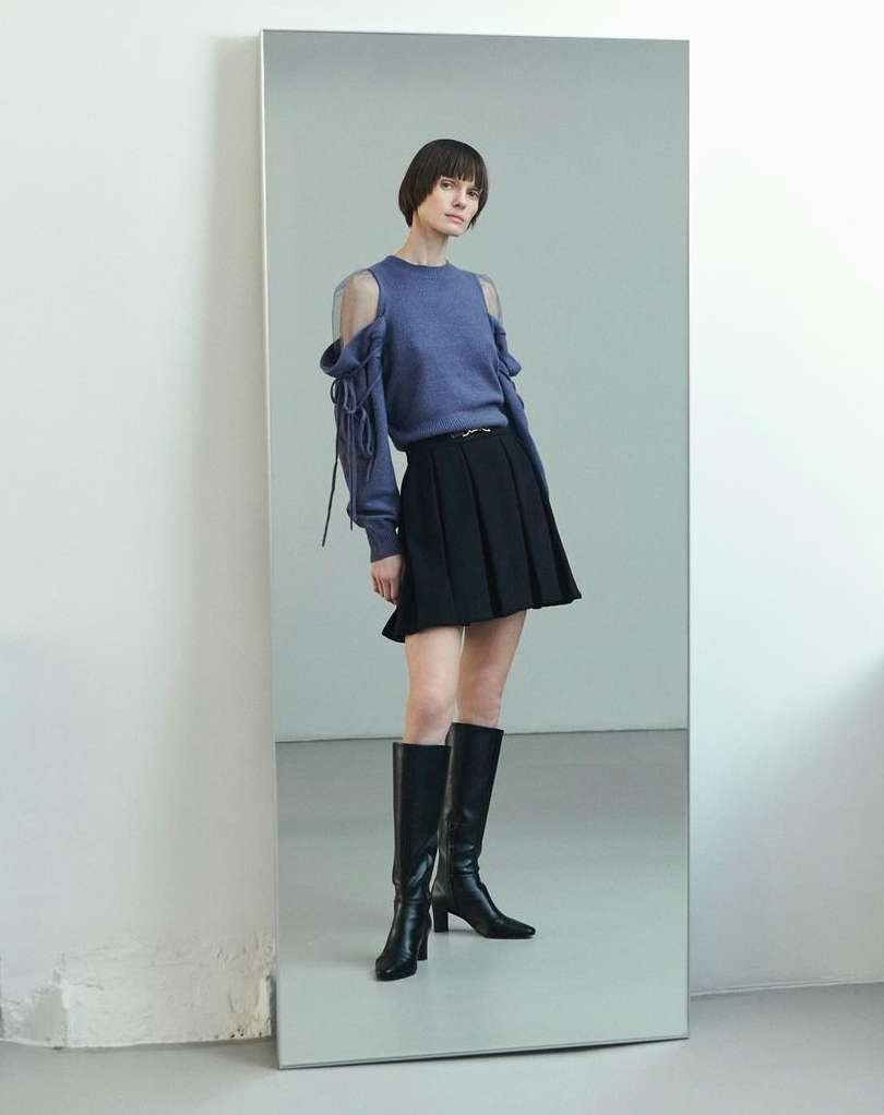 A slender woman with dark bobbed hair poses in a cut-out blue sweater, short black pleated skirt, and high black boots against a gray background.