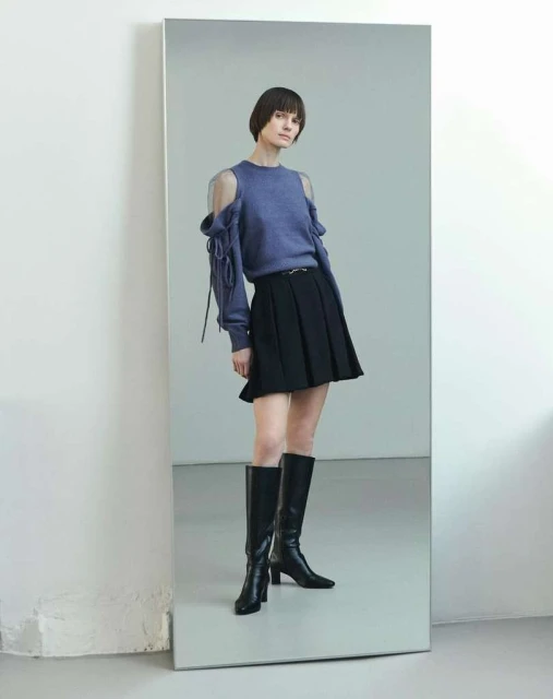 A slender woman with dark bobbed hair poses in a cut-out blue sweater, short black pleated skirt, and high black boots against a gray background.