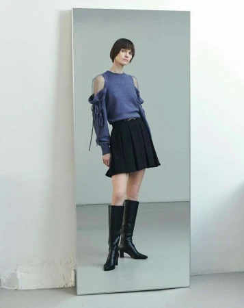 A slender woman with dark bobbed hair poses in a cut-out blue sweater, short black pleated skirt, and high black boots against a gray background.