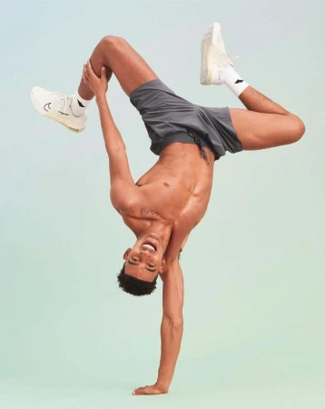 Shirtless athletic man in grey shorts doing a handstand pose against a gradient background.