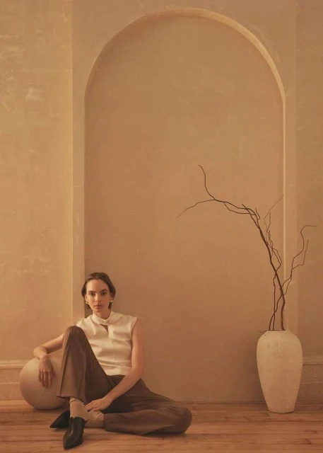 A young adult woman in neutral-toned clothing sitting on the floor next to a large round vase with bare branches in a minimalist room.