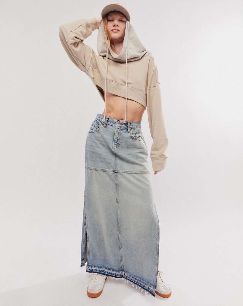 Young woman poses in a cropped hoodie, long denim skirt, and sneakers against a white backdrop in a studio setting with soft lighting.