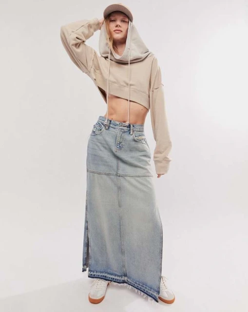 Young woman poses in a cropped hoodie, long denim skirt, and sneakers against a white backdrop in a studio setting with soft lighting.