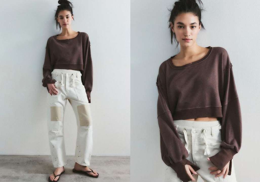 A woman poses in a brown long-sleeve crop top and cream-colored pants with a white wall background in a studio-style setting.
