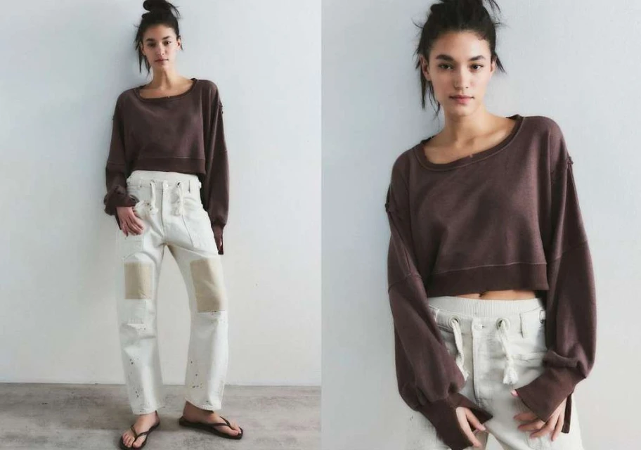 A woman poses in a brown long-sleeve crop top and cream-colored pants with a white wall background in a studio-style setting.