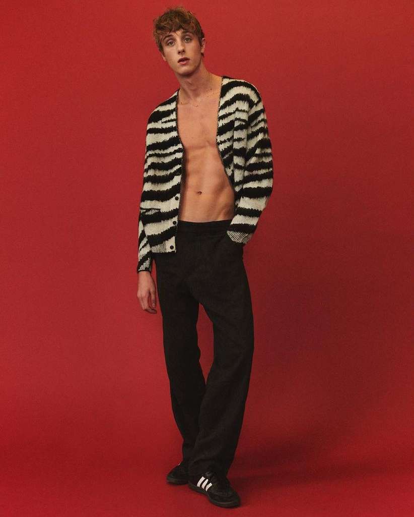 Man in black pants and a zebra-striped cardigan poses against a red backdrop, showcasing a modern fashion style with a studio vibe.
