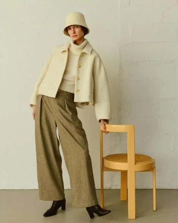 Model in cream bucket hat and jacket standing by yellow wooden chair against white brick wall