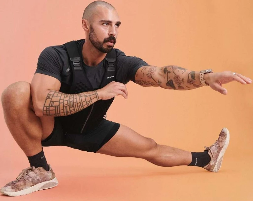 Adult male in athletic wear stretches legs in a studio, showing tattoos on his arms and legs against an orange background. 