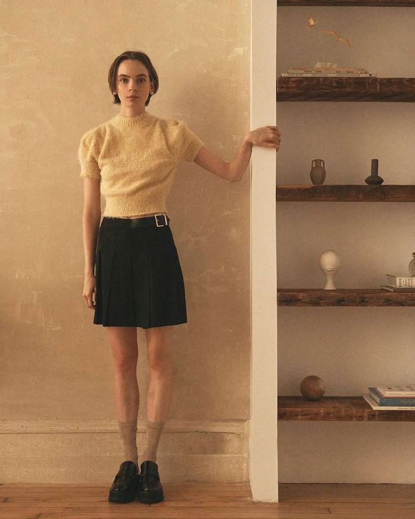 Full shot of a young adult female model in a soft yellow sweater and black skirt standing next to a wall and a bookshelf. Indoor, eye-level.