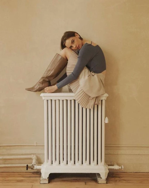 Adult woman in a grey top and light tan skirt, seated on top of a white radiator in an indoor setting with a beige wall background.