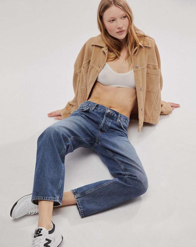 A young woman with red hair, wearing a light brown jacket and blue jeans, sits casually in a studio setting with natural lighting.