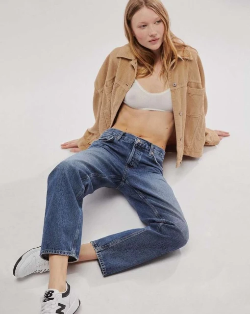 A young woman with red hair, wearing a light brown jacket and blue jeans, sits casually in a studio setting with natural lighting.