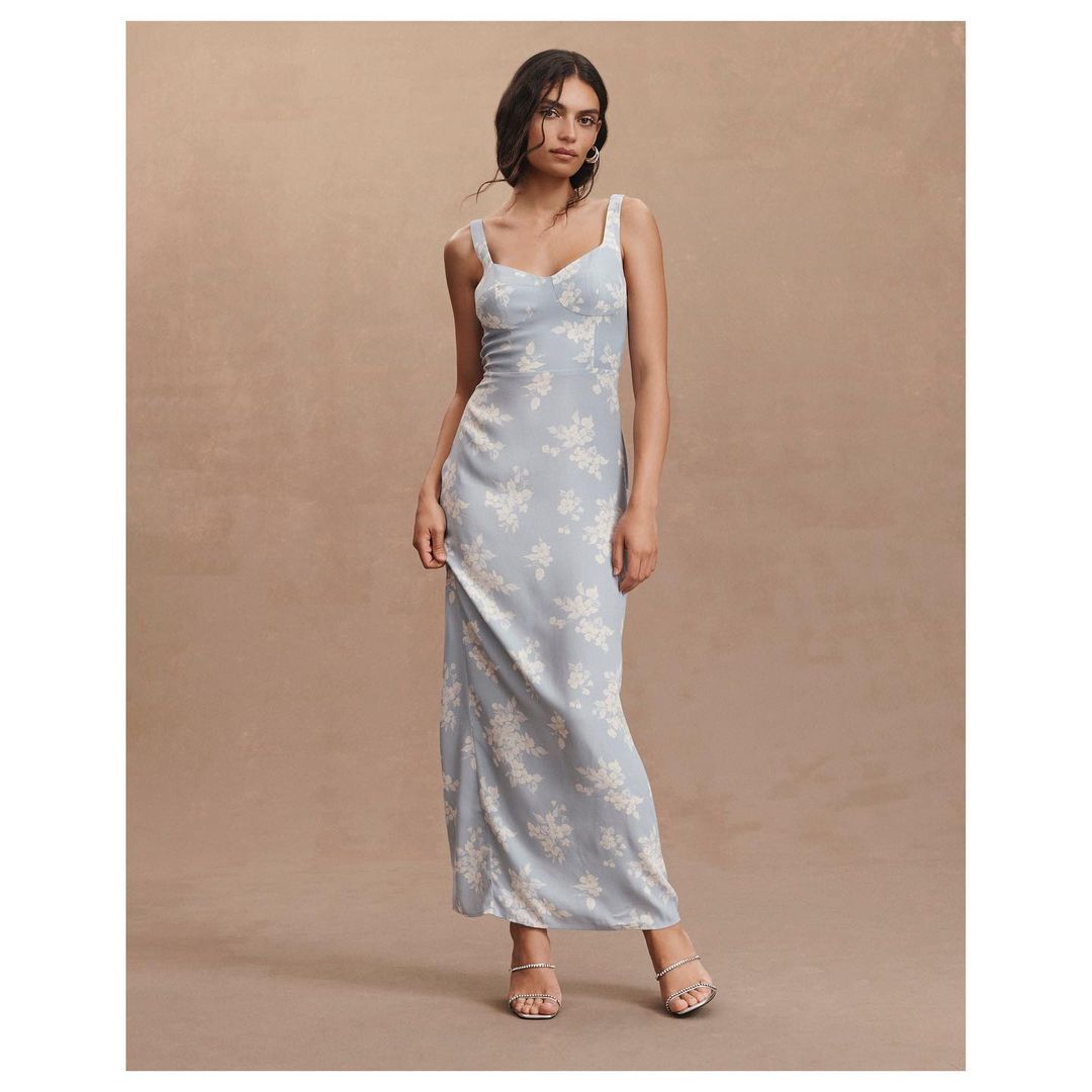 An adult female fashion model wears a pale blue floral maxi dress with spaghetti straps in a neutral, light-filled studio setting.