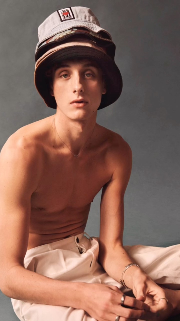 Young adult male with bare chest poses with three bucket hats stacked on his head in a portrait against a grey backdrop.
