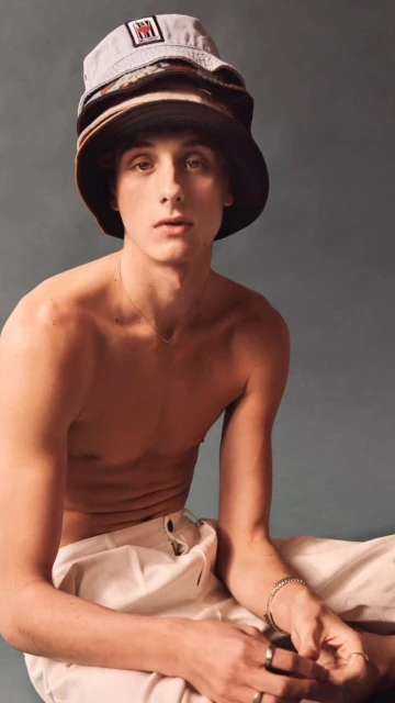 Young adult male with bare chest poses with three bucket hats stacked on his head in a portrait against a grey backdrop.