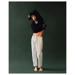 Young man with curly hair wearing a dark sweater pulled over his head and light grey trousers against a dark green studio backdrop.