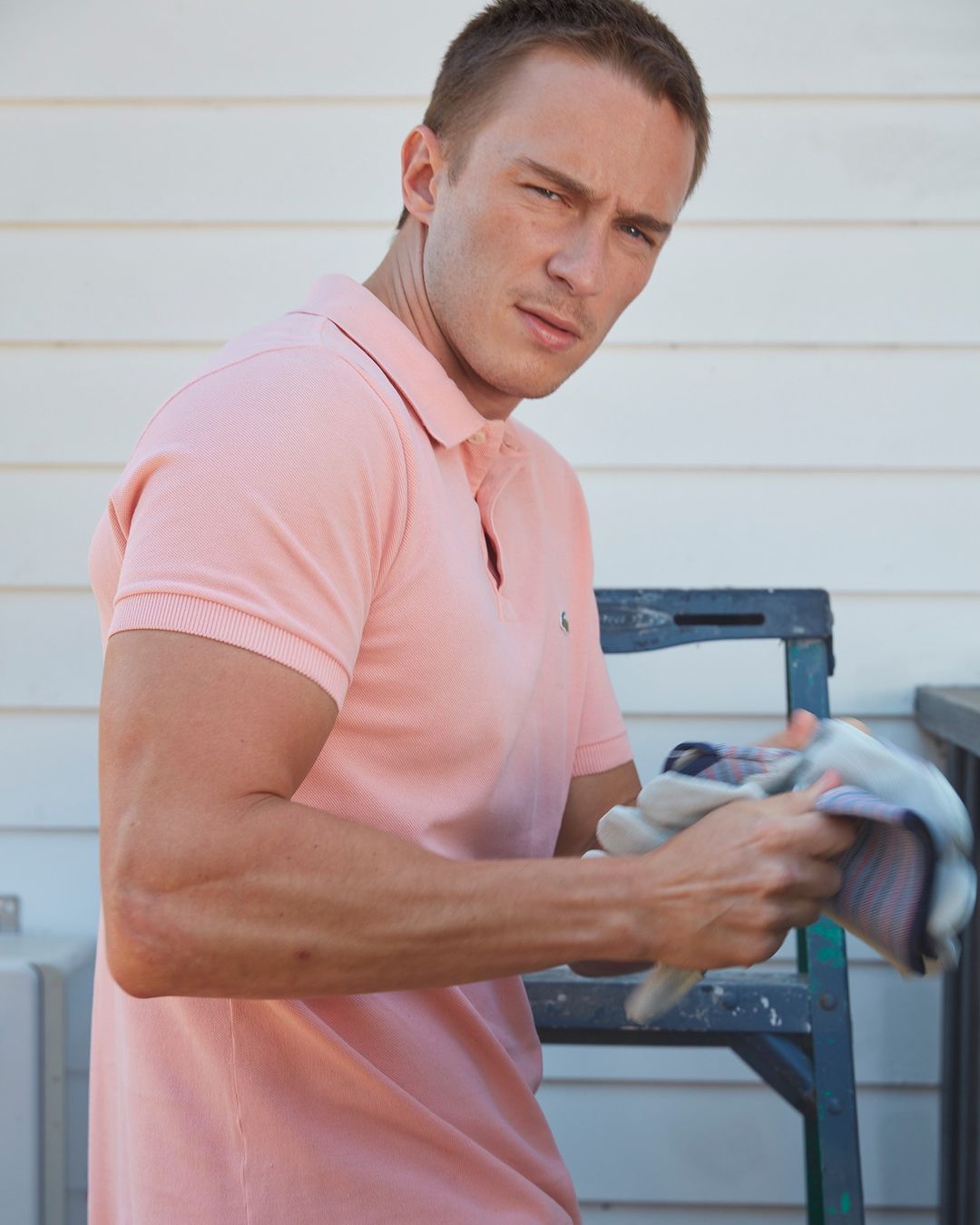 Adult male in a pink polo shirt stands near a ladder, wiping his hands with a cloth in an outdoor setting with natural lighting.