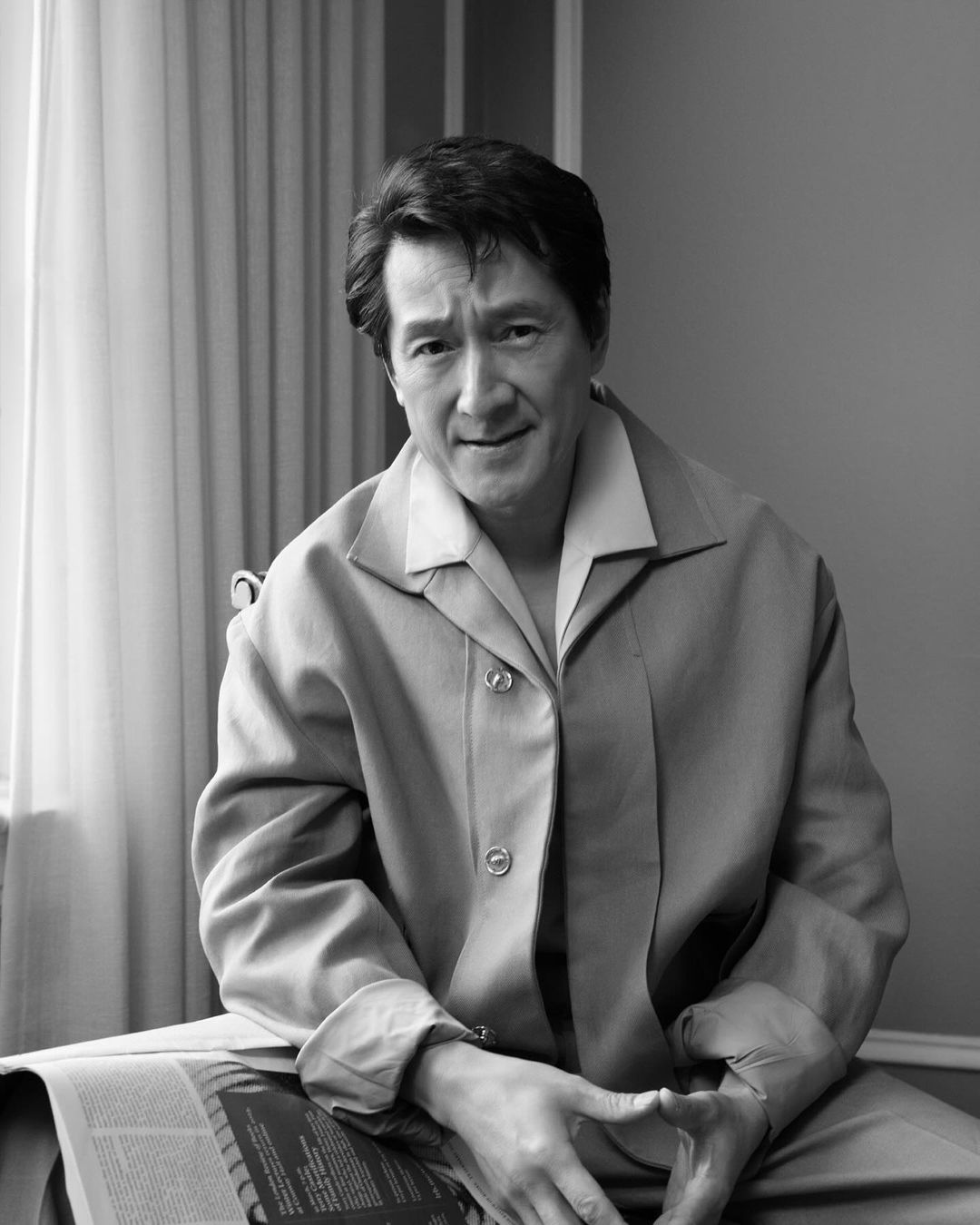 Black and white portrait of a mature adult man sitting indoors with a newspaper, wearing a collared shirt and button-down jacket.