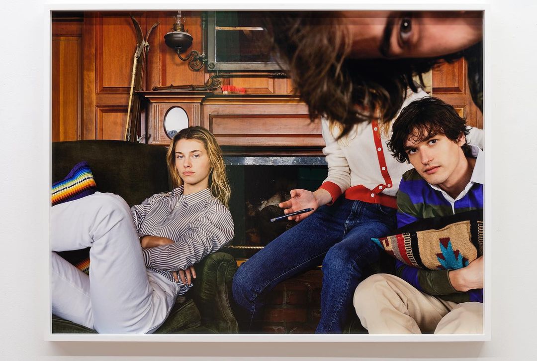 Three people pose indoors in front of wood-paneled wall: a seated woman in striped shirt, a seated woman and man in sweaters.