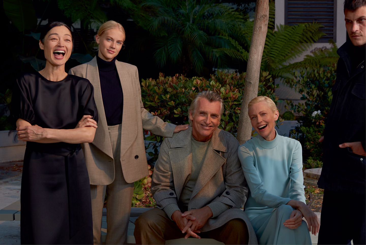 Group of five people, including three women and two men, posing outdoors with green foliage and trees visible in the background.