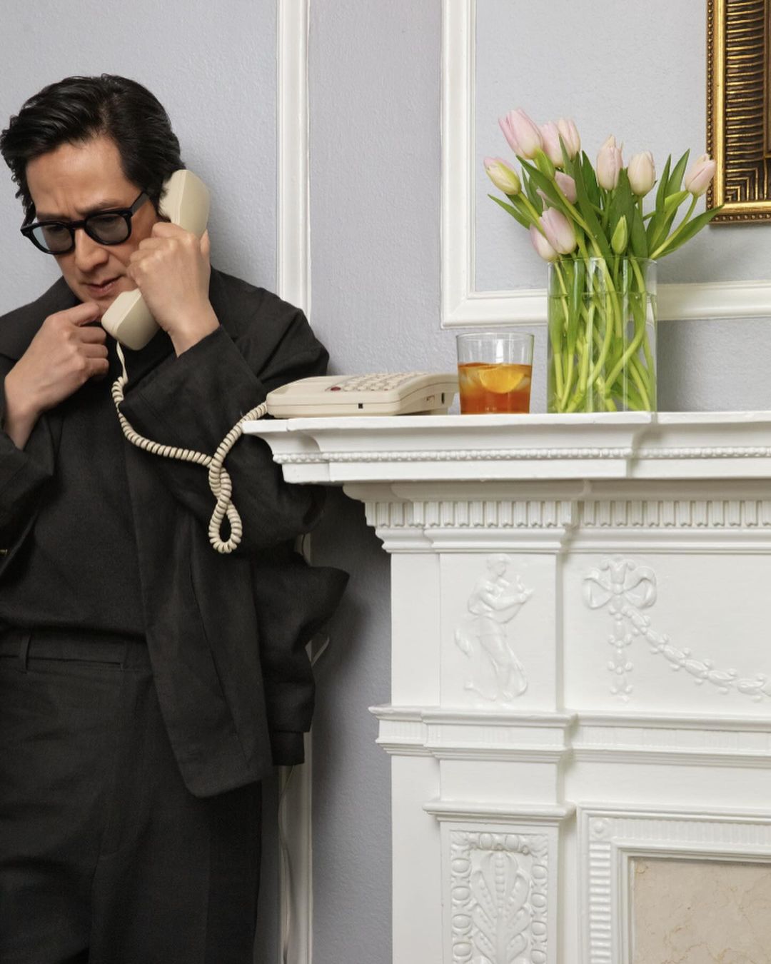 A man in a suit holds a landline telephone receiver up to his ear, standing next to a white fireplace mantle with tulips and a drink.