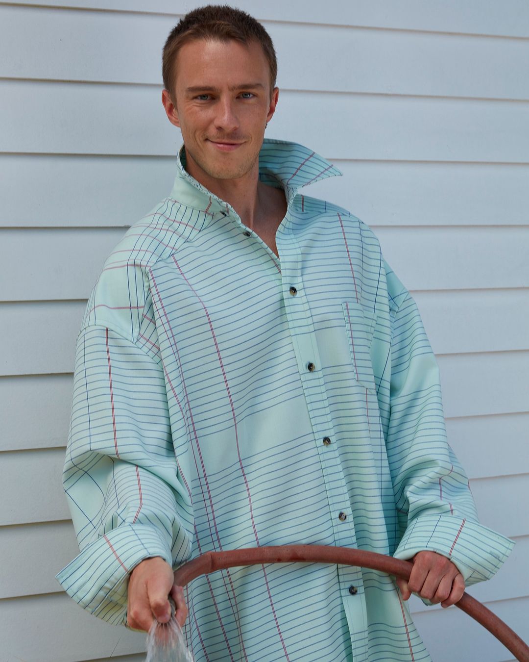 Smiling man in oversized plaid shirt poses outdoors holding a garden hose, water flowing. Youthful, casual fashion, daylight.