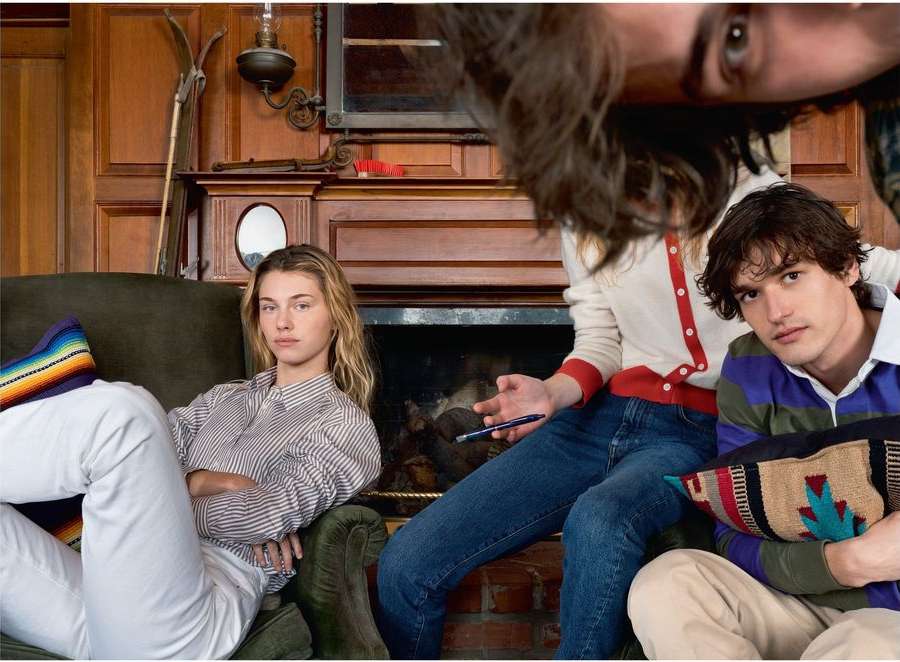 Three people in a vintage-style room with a fireplace, two seated and one partially visible peering down from above.