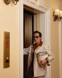 Man wearing sunglasses and a beige jacket exiting an elevator, holding an ice bucket in a hotel hallway.