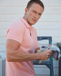Man wearing a pink polo shirt holding work gloves near a dark ladder and light siding