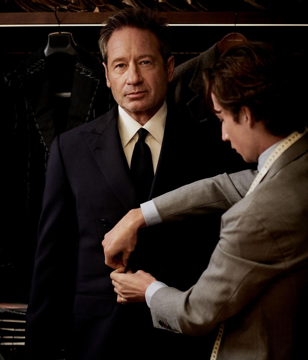 David Duchovny getting a suit tailored by a tailor in a suit shop, portrait shot, clothing rack in the background.