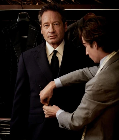 David Duchovny getting a suit tailored by a tailor in a suit shop, portrait shot, clothing rack in the background.