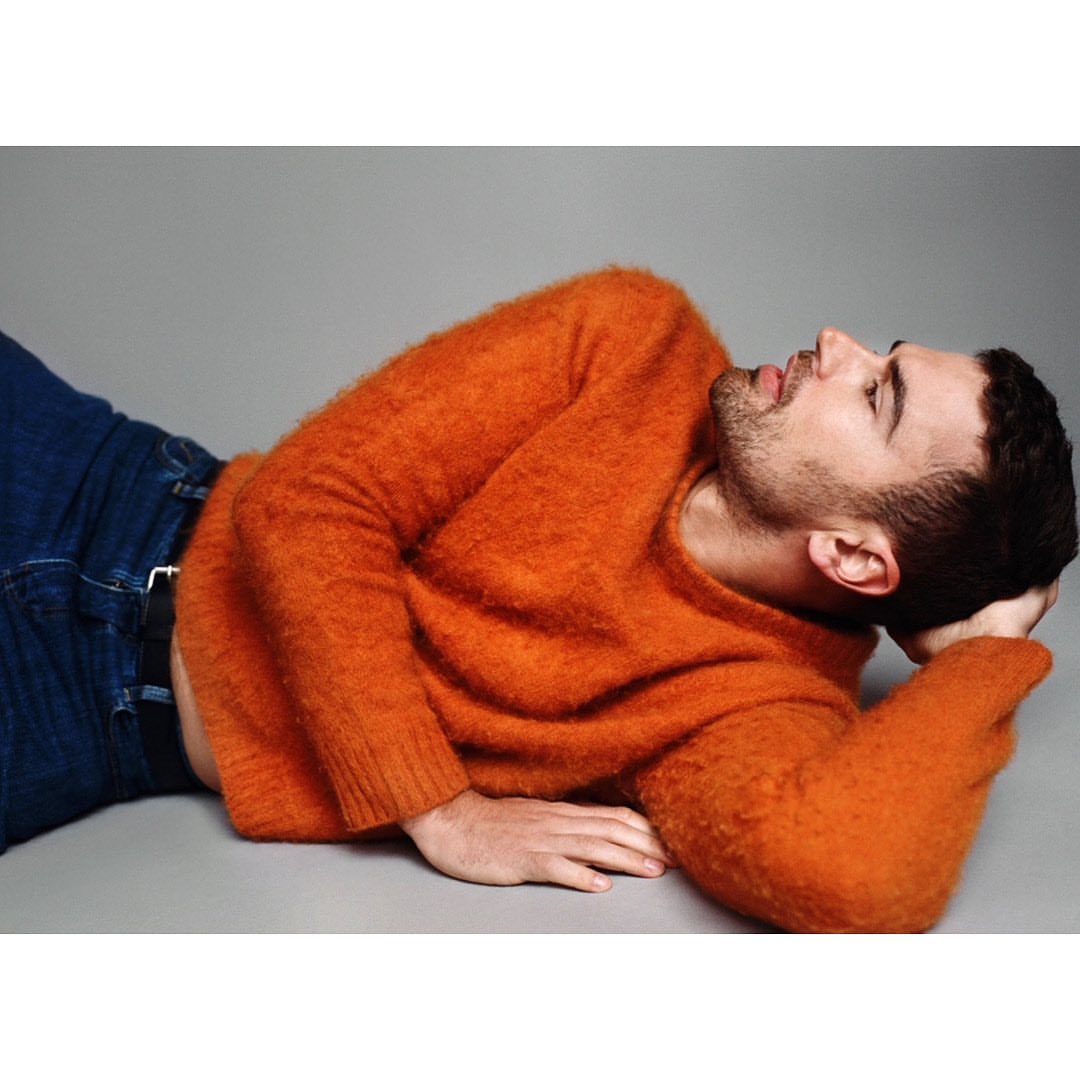 Man lying down in an orange sweater and jeans in a studio. He is resting his head on his hand and looking up and to the right.