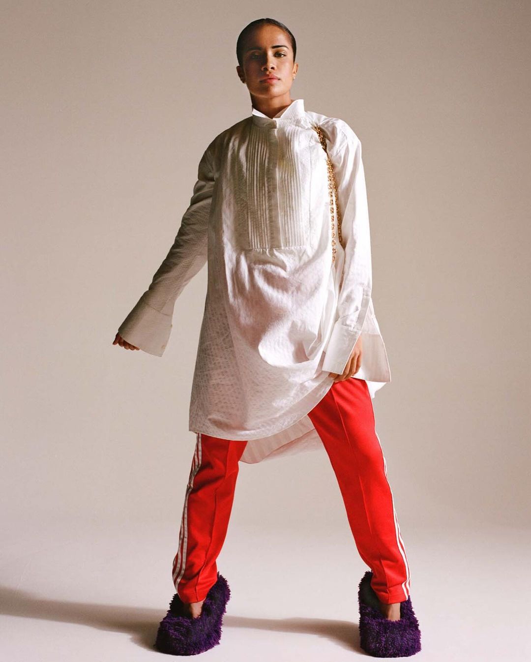Woman in an oversized white shirt, red striped pants, and fluffy slippers stands in a photo studio with a neutral background.