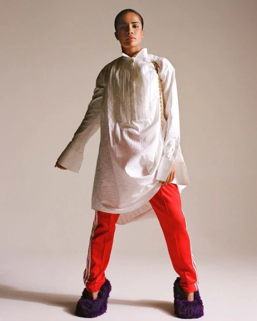 Woman in an oversized white shirt, red striped pants, and fluffy slippers stands in a photo studio with a neutral background.