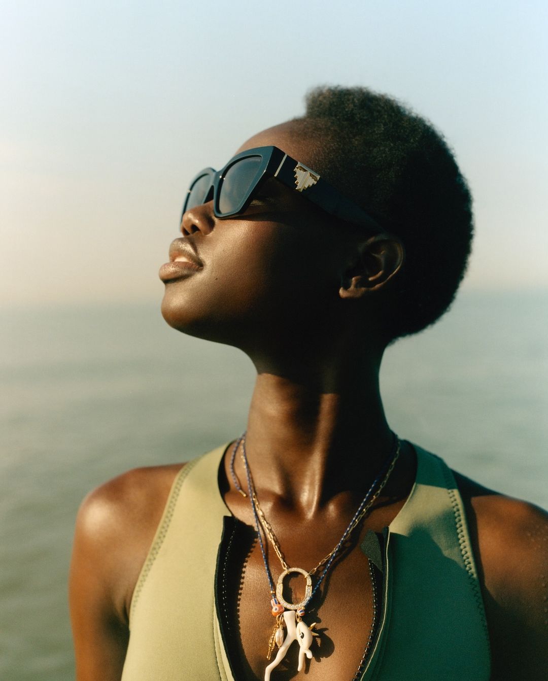 An adult woman with short hair and sunglasses poses outdoors wearing a swimsuit top and necklaces against a blurred ocean background.