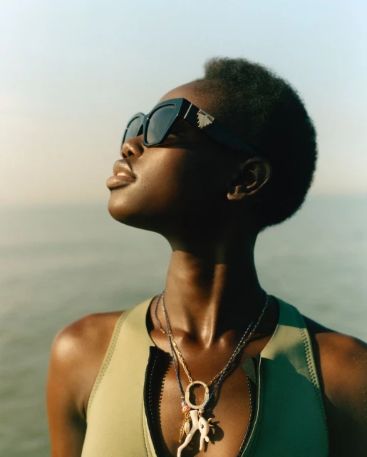An adult woman with short hair and sunglasses poses outdoors wearing a swimsuit top and necklaces against a blurred ocean background.