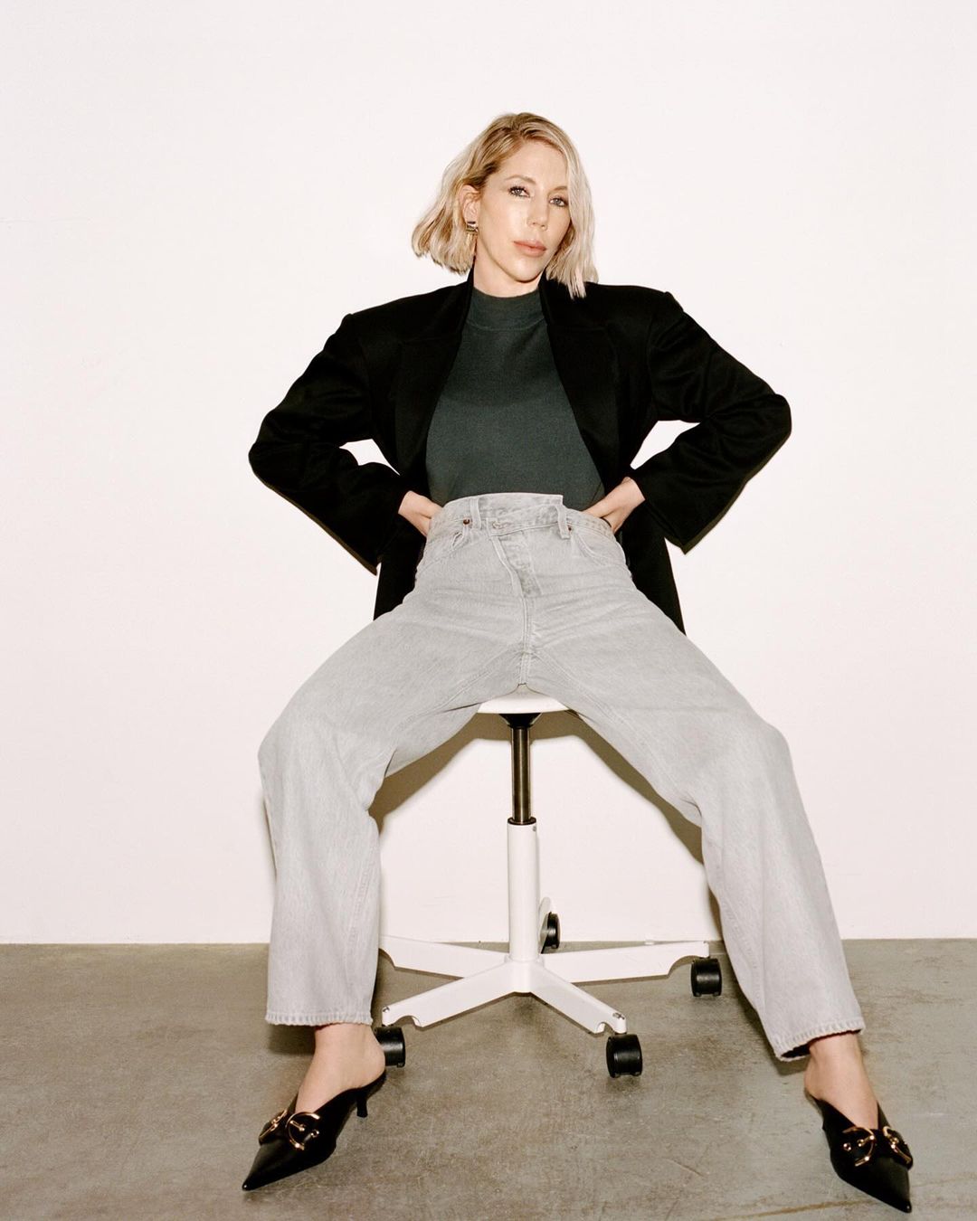 A blonde woman in a black blazer and light-wash jeans sits on a white rolling chair against a white wall in a casual fashion shot.