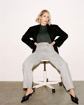 A blonde woman in a black blazer and light-wash jeans sits on a white rolling chair against a white wall in a casual fashion shot.