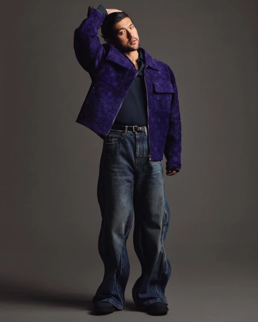 A full-body shot of a young adult man with dark hair, wearing a purple jacket and blue jeans, posing in a photo studio with a grey background.