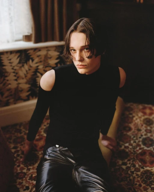 Portrait of a young adult with dark hair wearing a black, cut-out shoulder top and dark shiny pants sitting on a patterned carpet indoors.