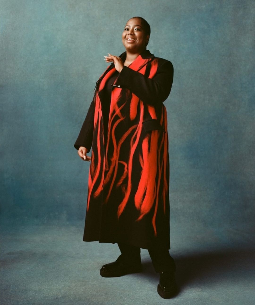 Full-length portrait of an adult woman in a patterned black and red coat against a mottled blue background in a studio setting.