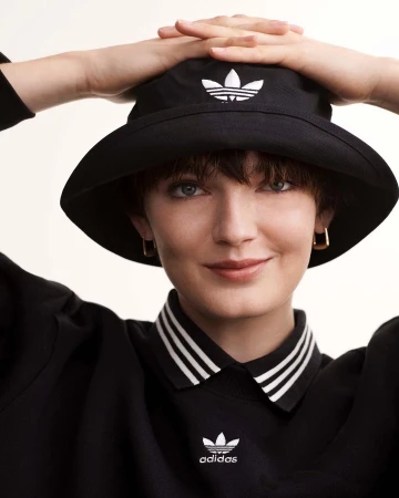 Close-up portrait of a woman wearing a black bucket hat and black collared shirt with white stripes against a plain background