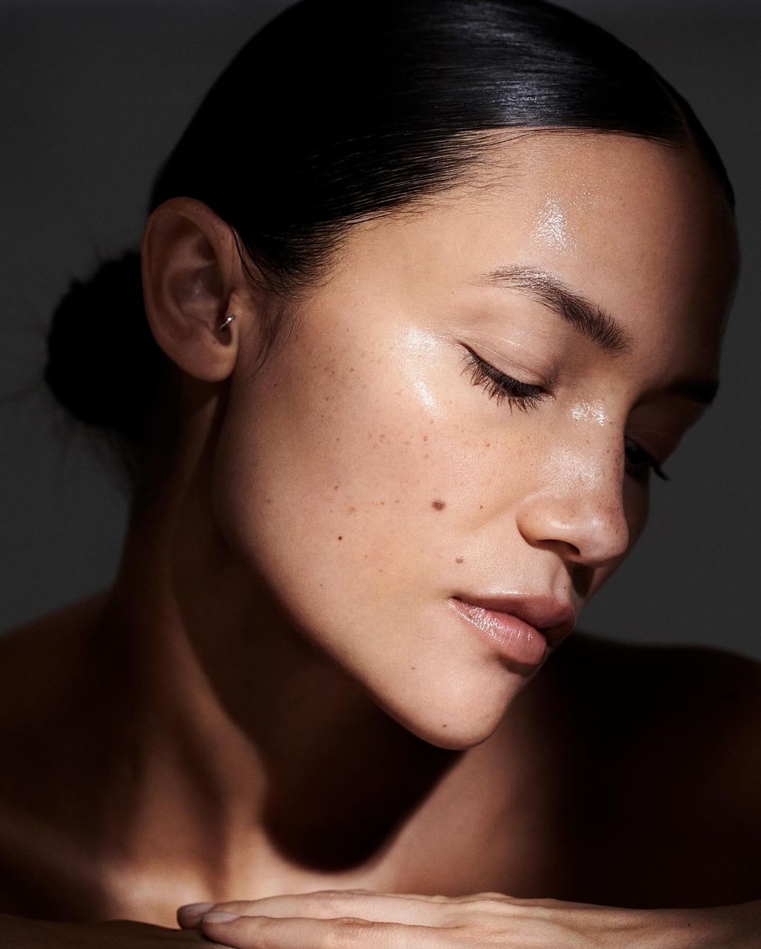Close-up beauty shot of a woman with her eyes closed, fair skin, freckles, and dark hair pulled back, against a dark grey background.