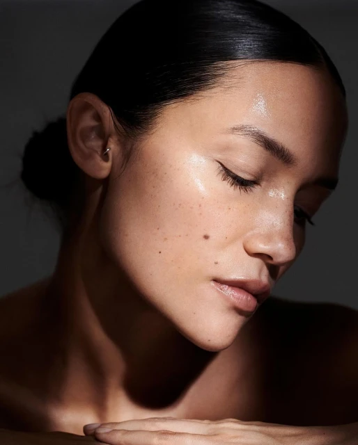 Close-up beauty shot of a woman with her eyes closed, fair skin, freckles, and dark hair pulled back, against a dark grey background.