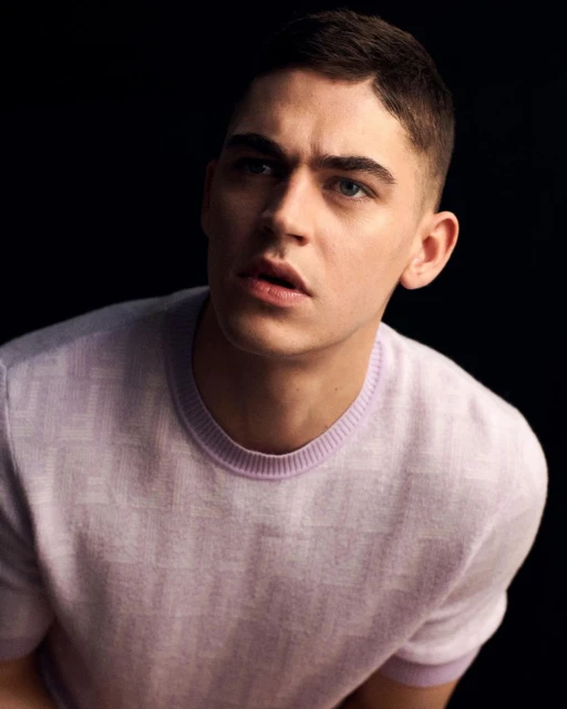 Portrait of a young man with short brown hair wearing a lavender patterned sweater against a dark background, gazing upwards.