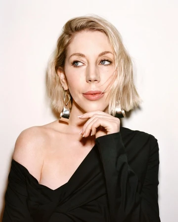 Portrait of a blonde woman wearing a black off-the-shoulder top and sculptural silver earrings against a plain white background.