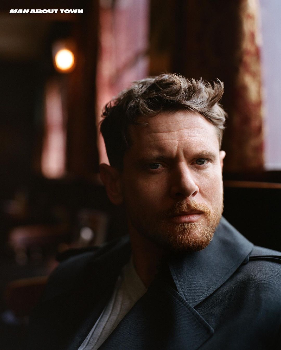 Portrait of an adult male with a beard wearing a coat, posing indoors in front of a partially visible window with a light fixture.